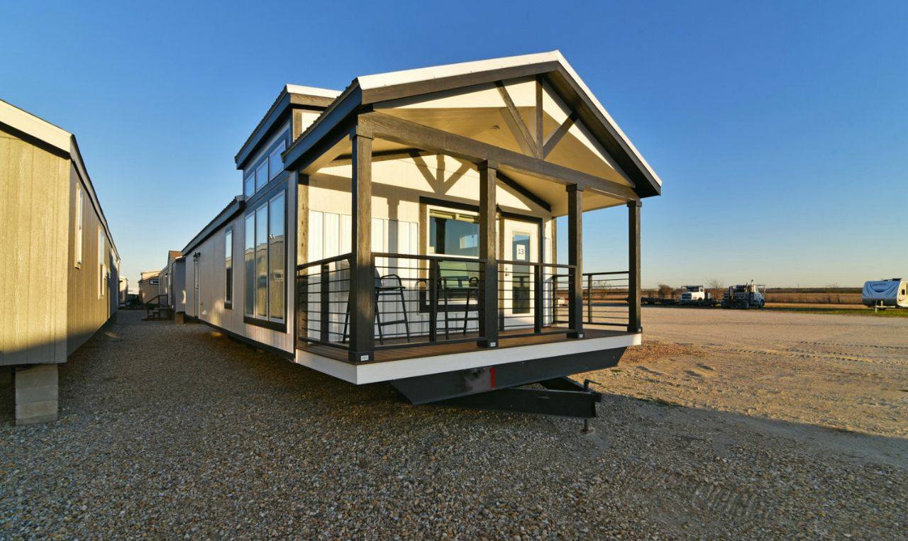 A modern manufactured home with a pitched roof and large windows. Sunlit porch with chairs, set on gravel under a clear blue sky, evokes tranquility.