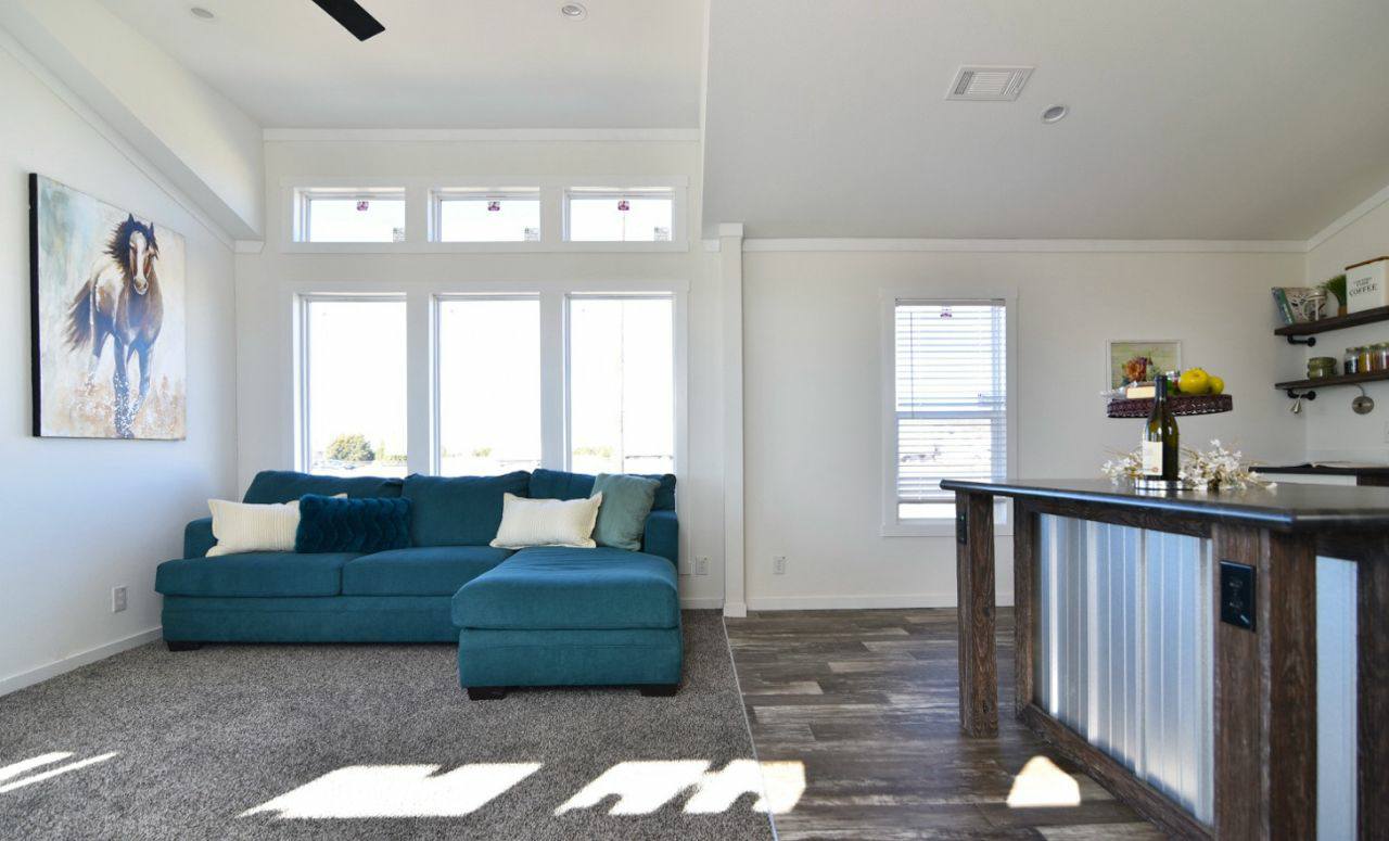 A bright living area with a teal sectional sofa on gray carpet, a horse painting on the wall, and a bar with wooden accents. Large windows and a serene ambiance.