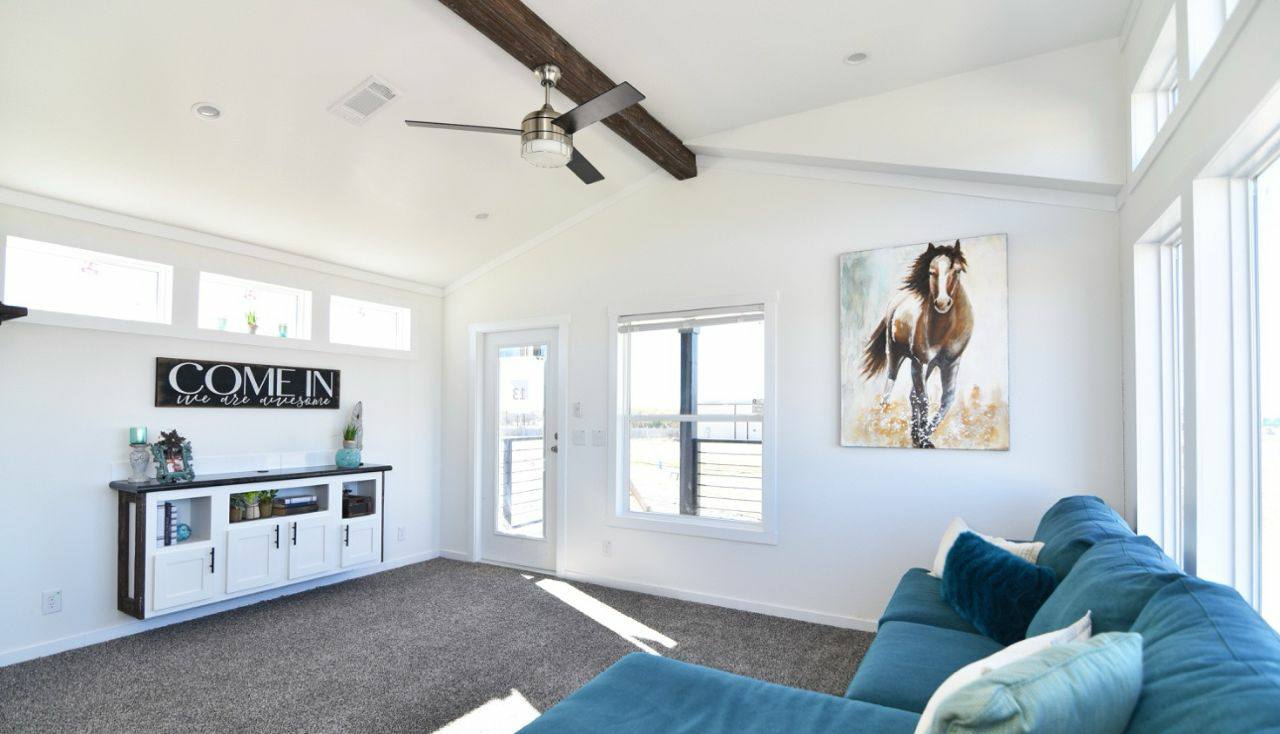 Modern, bright living room with white walls and vaulted ceiling featuring wood beams. There's a blue sectional sofa, horse painting, and a sideboard with a sign reading “COME IN."