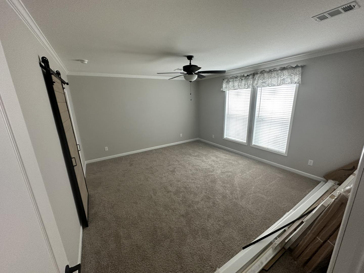 Empty room with light gray walls, beige carpet, and a ceiling fan. A barn door is on the left, and two large windows with curtains on the right.