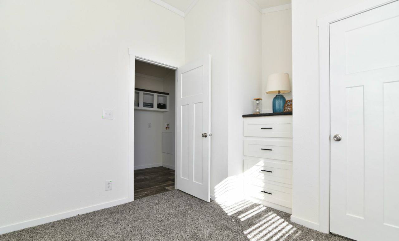 Minimalist room with white walls and carpeted floor. Features a white dresser, a blue vase, and a lamp. Light streams through blinds, creating shadows. Cozy ambiance.