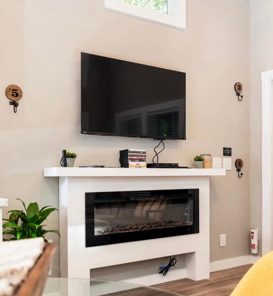 Modern living room with a mounted flat-screen TV above a sleek white electric fireplace. Minimalist decor with small potted plants, DVDs, and neutral tones.