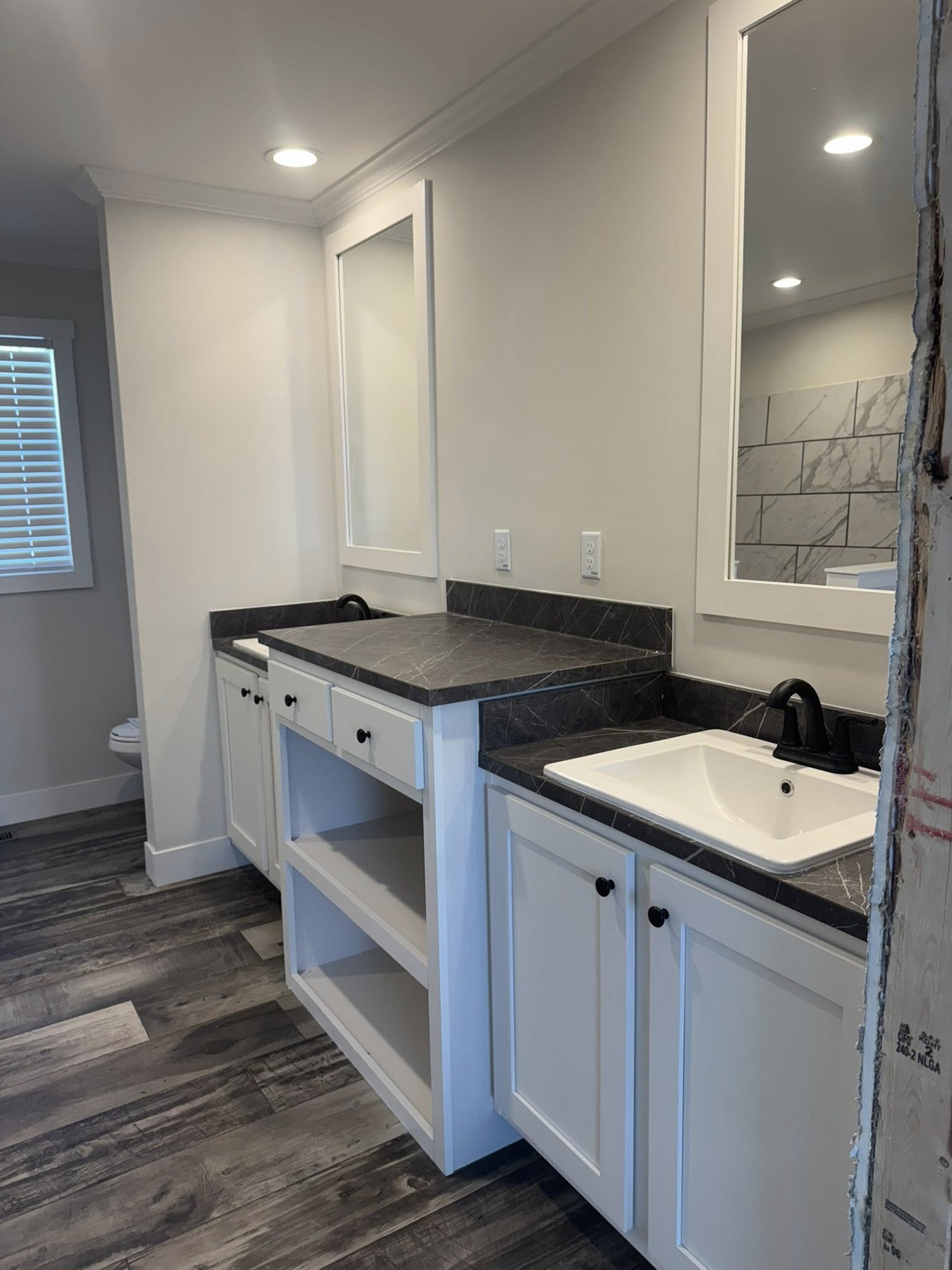 Modern bathroom with dark wood flooring and recessed lighting. Features a white double vanity with black fixtures, gray countertops, and two mirrors.