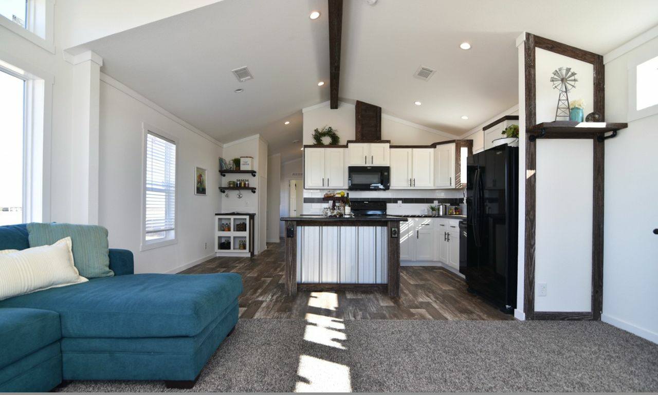 Bright, modern living area with vaulted ceiling, featuring a teal sectional sofa and striped cushions. Open kitchen with white cabinets and dark accents, creating an inviting and cozy atmosphere.