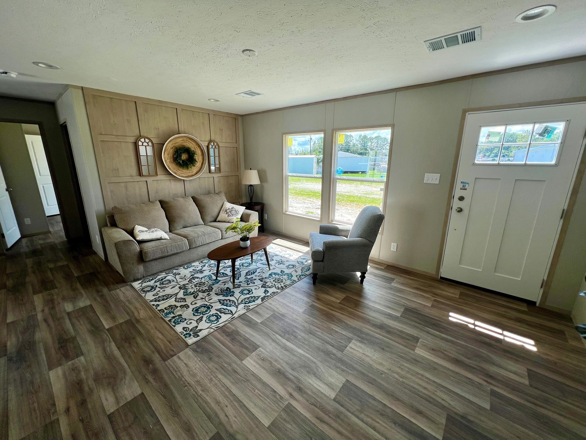 Cozy living room with wood flooring, featuring a beige couch, patterned rug, and gray armchair. Sunlight streams through large windows, creating a warm ambiance.