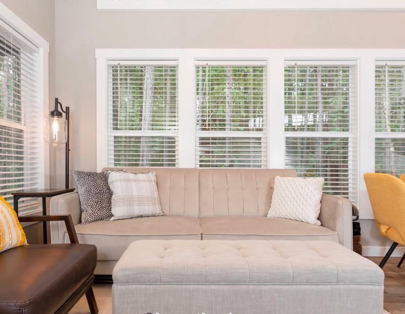 Inviting living room with beige sofa, three patterned cushions, and tufted ottoman. Large windows with blinds offer a serene view of trees. Bright and cozy ambiance.