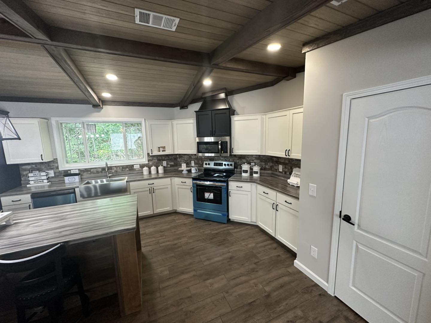 Spacious kitchen with rustic wood ceiling, white cabinets, and a blue stove. Large window provides natural light, highlighting the warm, inviting ambiance.