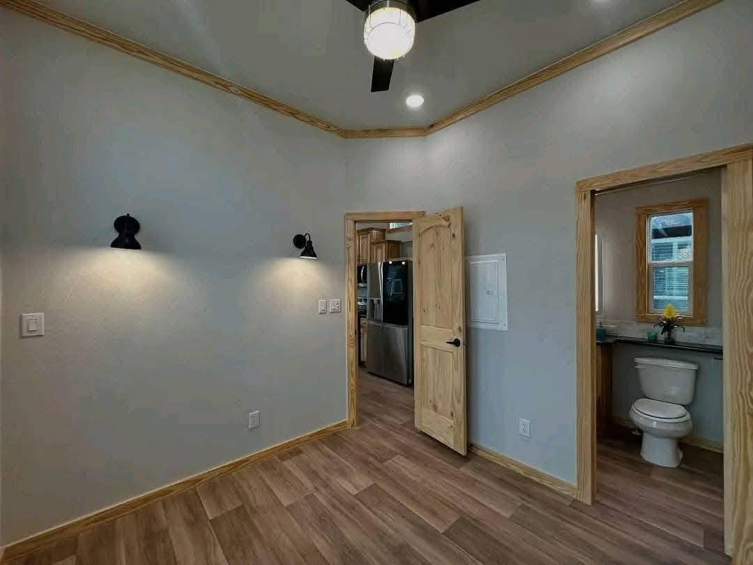 A room with light gray walls, wood trim, and laminate flooring. To the right, a bathroom with a toilet is visible. A partially open door reveals a kitchen with stainless steel appliances. Warm wall sconces illuminate the space, creating a cozy atmosphere.