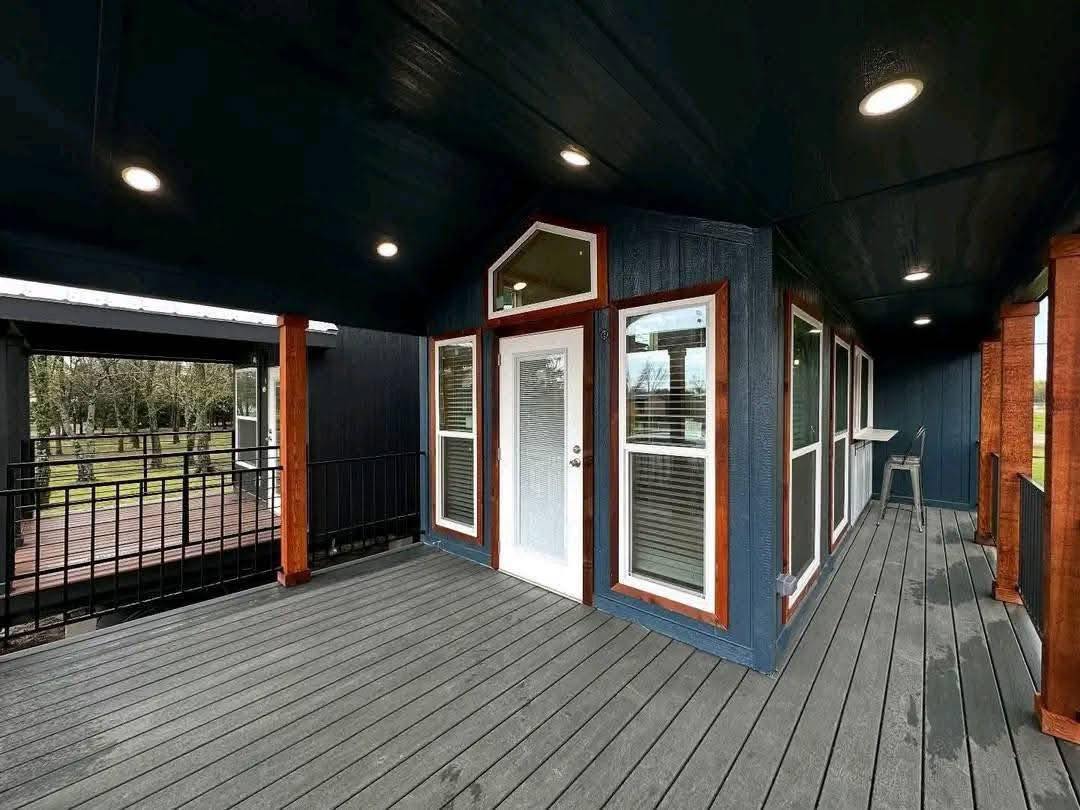 Covered porch with dark wood decking, white-trimmed glass doors and windows against blue walls. Ceiling lights create a welcoming ambiance. Scenic background.
