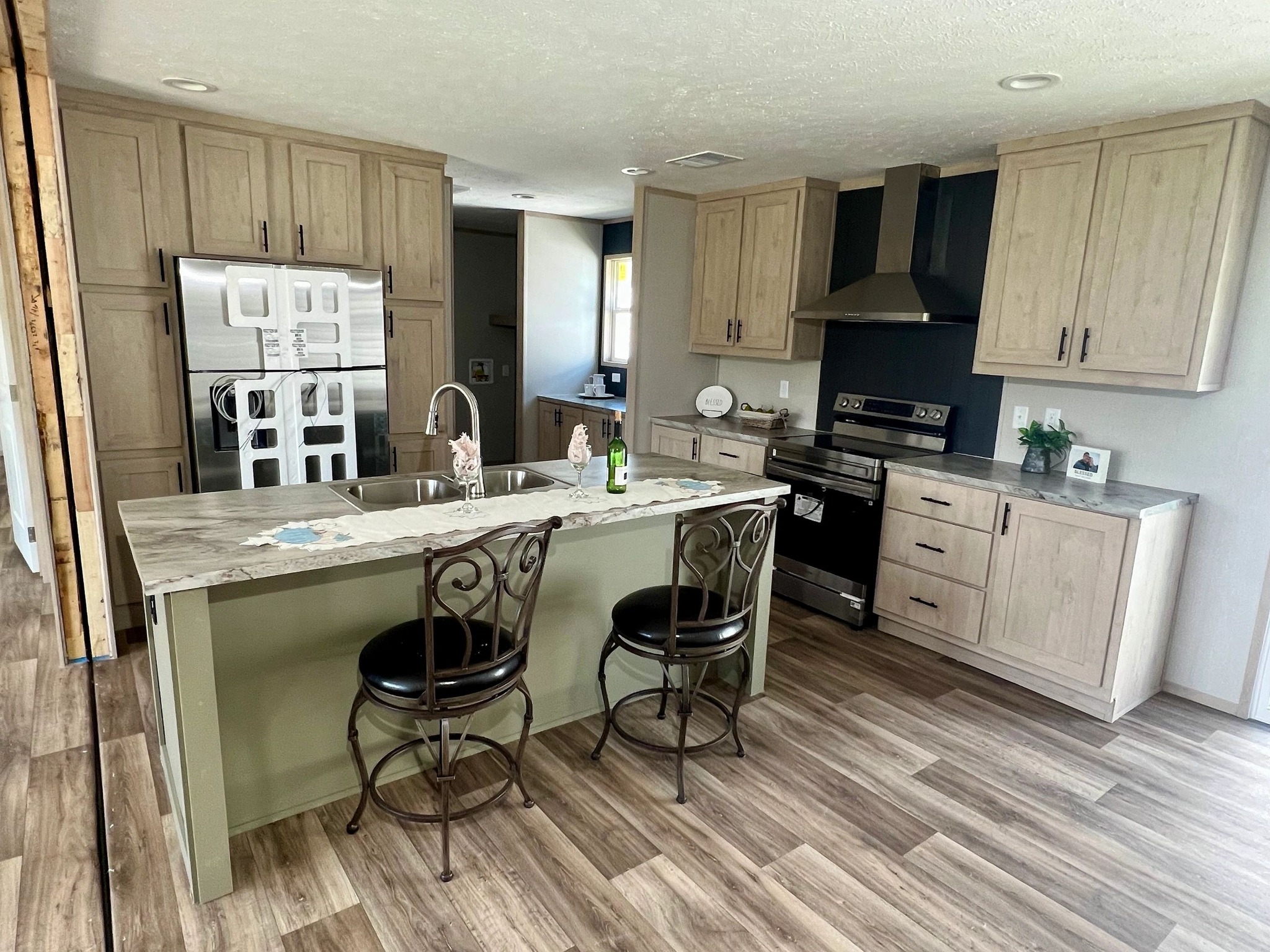 Spacious kitchen with light wood cabinets, stainless steel appliances including a fridge and oven, and an island with two ornate stools. Bright, modern feel.