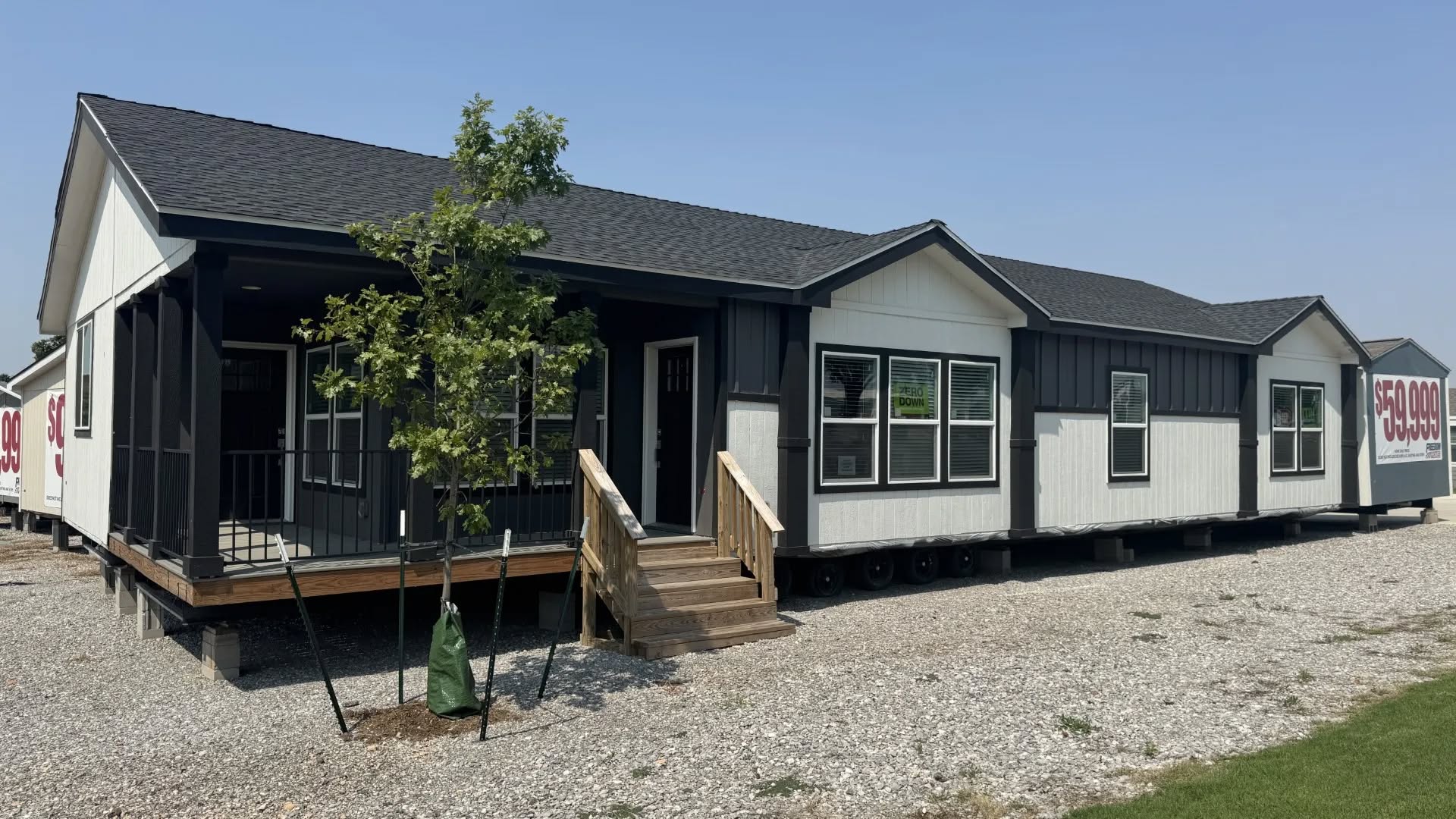 A modern modular home with a dark roof and white siding, featuring a small porch and staircase. The price, $59,990, is displayed on the side. A tree adds greenery.