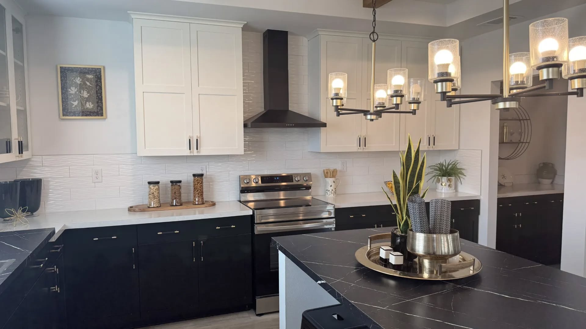Modern kitchen with black and white cabinets, stainless steel appliances, and a central island. A chandelier hangs above, adding elegance. The decor includes plants and neatly arranged canisters, creating a sleek, inviting atmosphere.