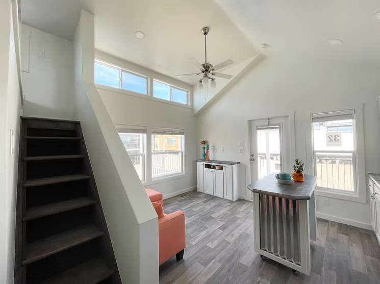 Modern, bright living room with white walls, vaulted ceiling, dark wood floors. Features orange armchairs, a central island, and a staircase.