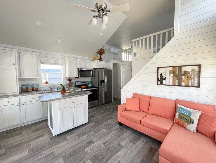 Modern tiny home interior with white cabinets and stainless-steel appliances. Coral sofa with "Live Laugh" pillow, and rustic decor creates a cozy atmosphere.