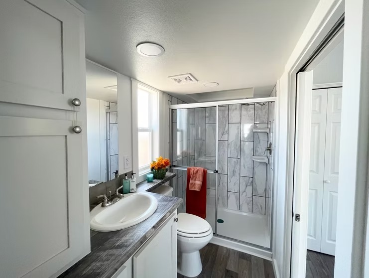 Modern bathroom with gray tile shower, white sink, and wooden countertop. Bright light from window, orange flowers, and red towel add warmth.