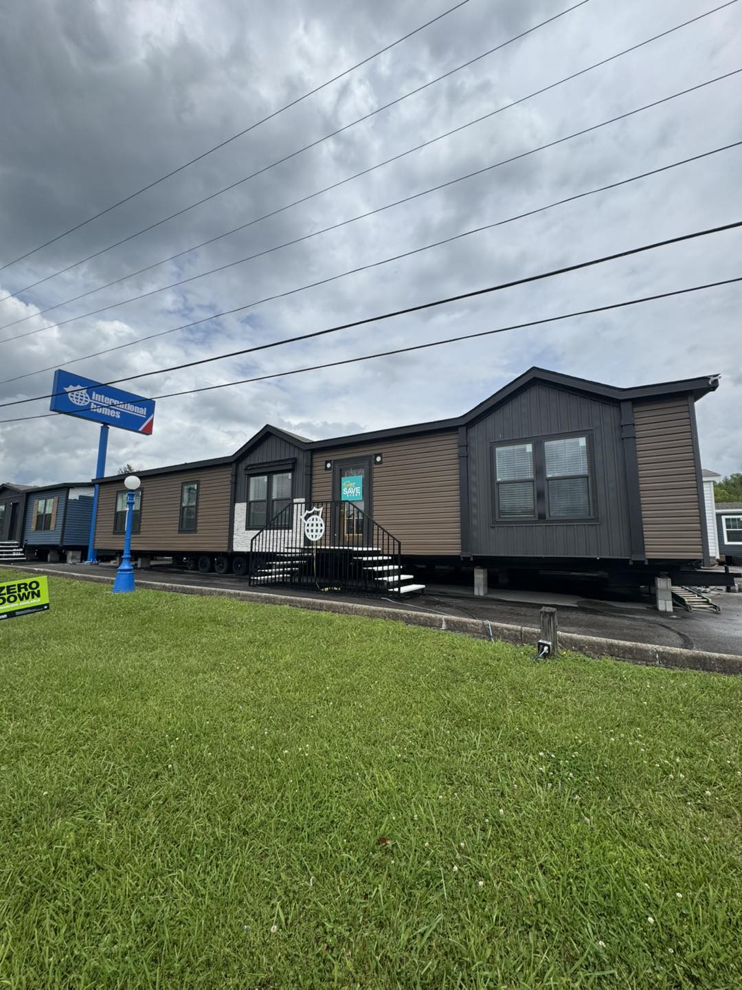 A large brown and gray mobile home is displayed on a lot with a green lawn, under a cloudy sky. A sign reads "Zero Down" and another promotes manufactured homes.