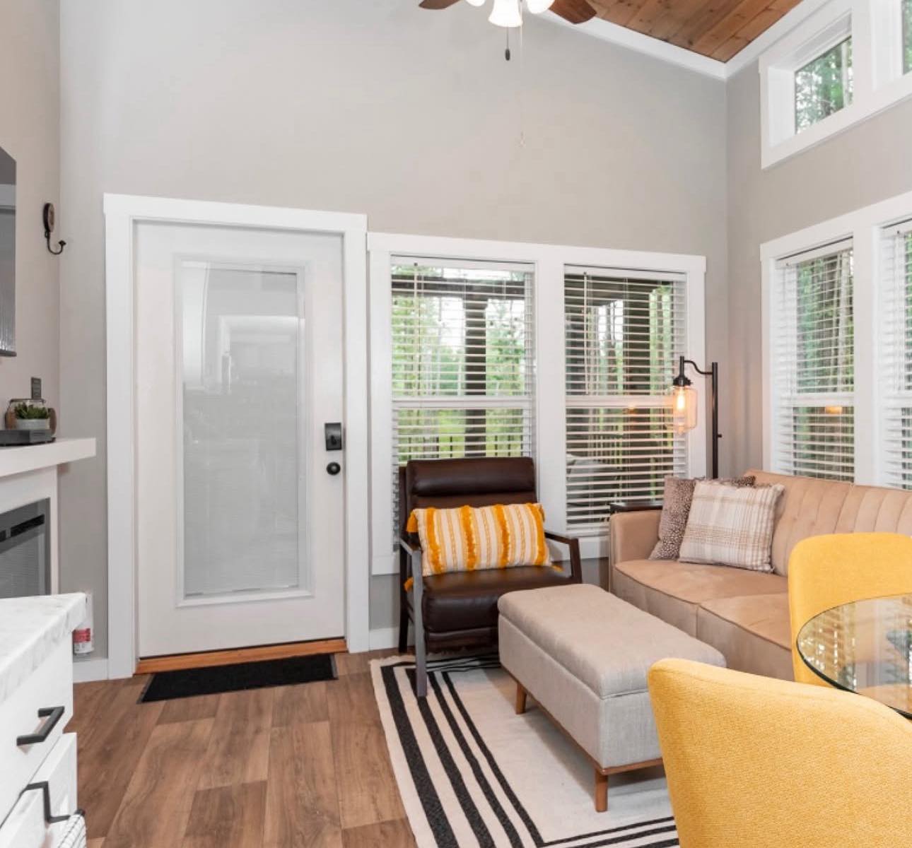 Cozy living room with natural light features a beige sectional, yellow pillows, striped rug, and glass table. Modern decor creates a welcoming atmosphere.