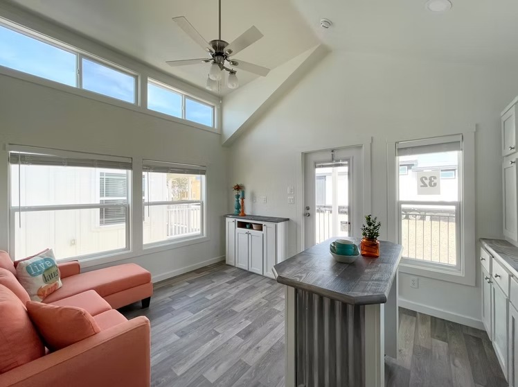 Bright living room with high ceiling and large windows. Features a peach sofa with colorful cushions, a ceiling fan, and a small kitchen island with decor.