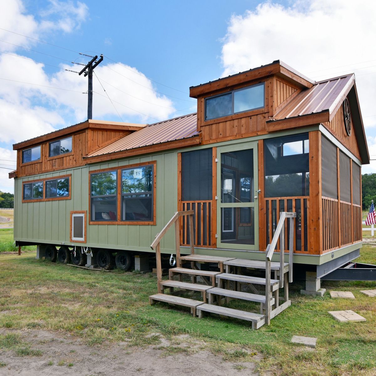 A rustic tiny house on wheels with a wooden exterior, large windows, and a small porch with stairs. It sits on grassy ground under a partially cloudy sky.