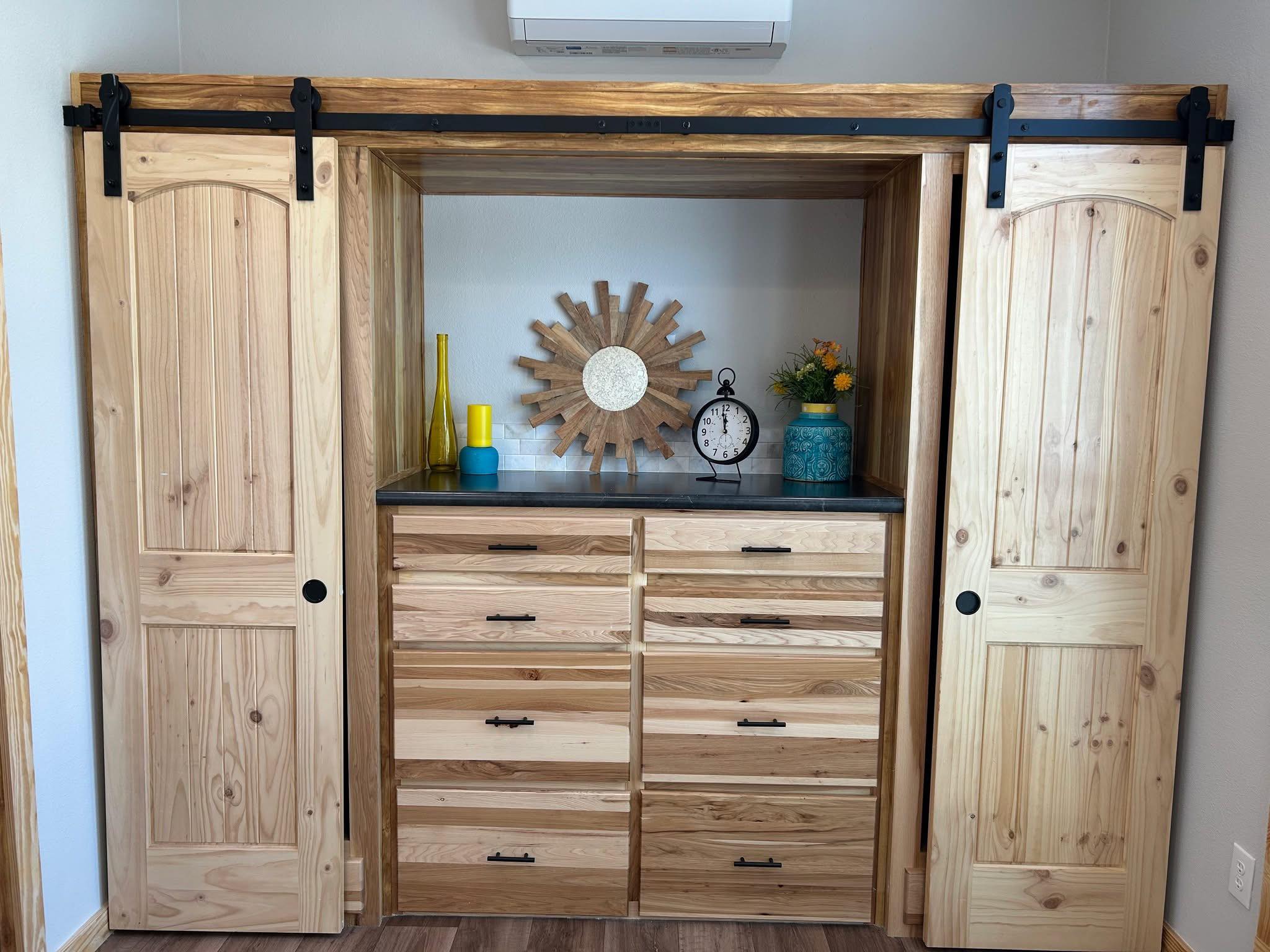 Built-in wooden dresser between sliding barn doors, topped with a sun-shaped mirror, clock, yellow vase, and blue jar with flowers, against a light wall.