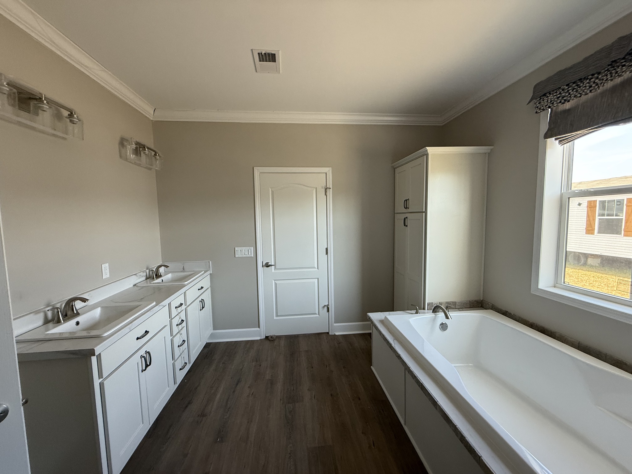Modern bathroom with white double sink vanity on the left, a large bathtub on the right, and a window. Light gray walls and wood flooring create a serene atmosphere.