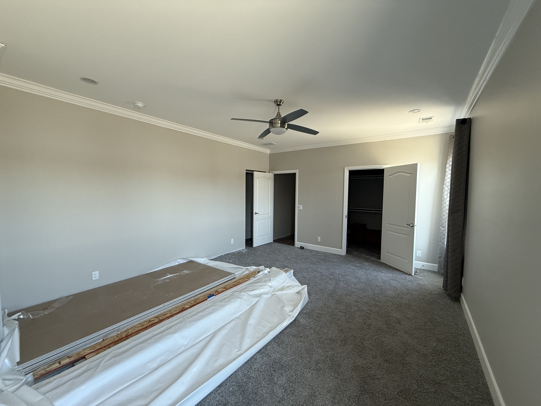 Empty room with gray walls, carpet, and ceiling fan. Two open doors lead to closets. Rolled-up white material and boards lie on the floor, hinting at ongoing setup.