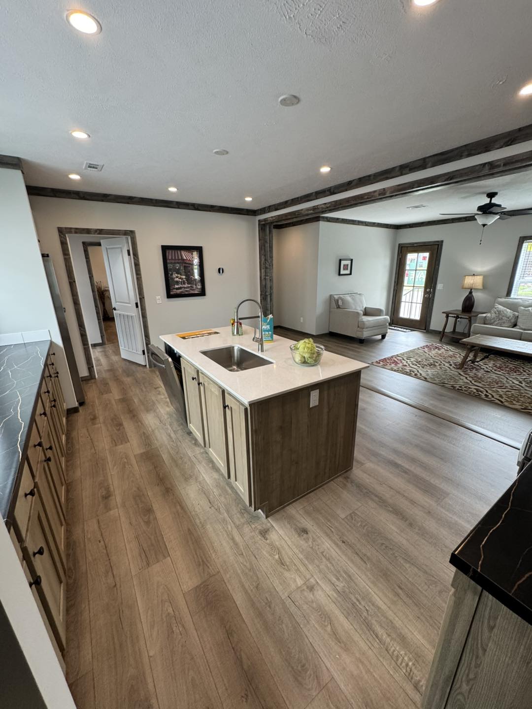 Modern kitchen and living area with wooden flooring. The kitchen features a central island with a sink and green apple centerpiece. A cozy living room with beige sofas and a rug is visible in the background.