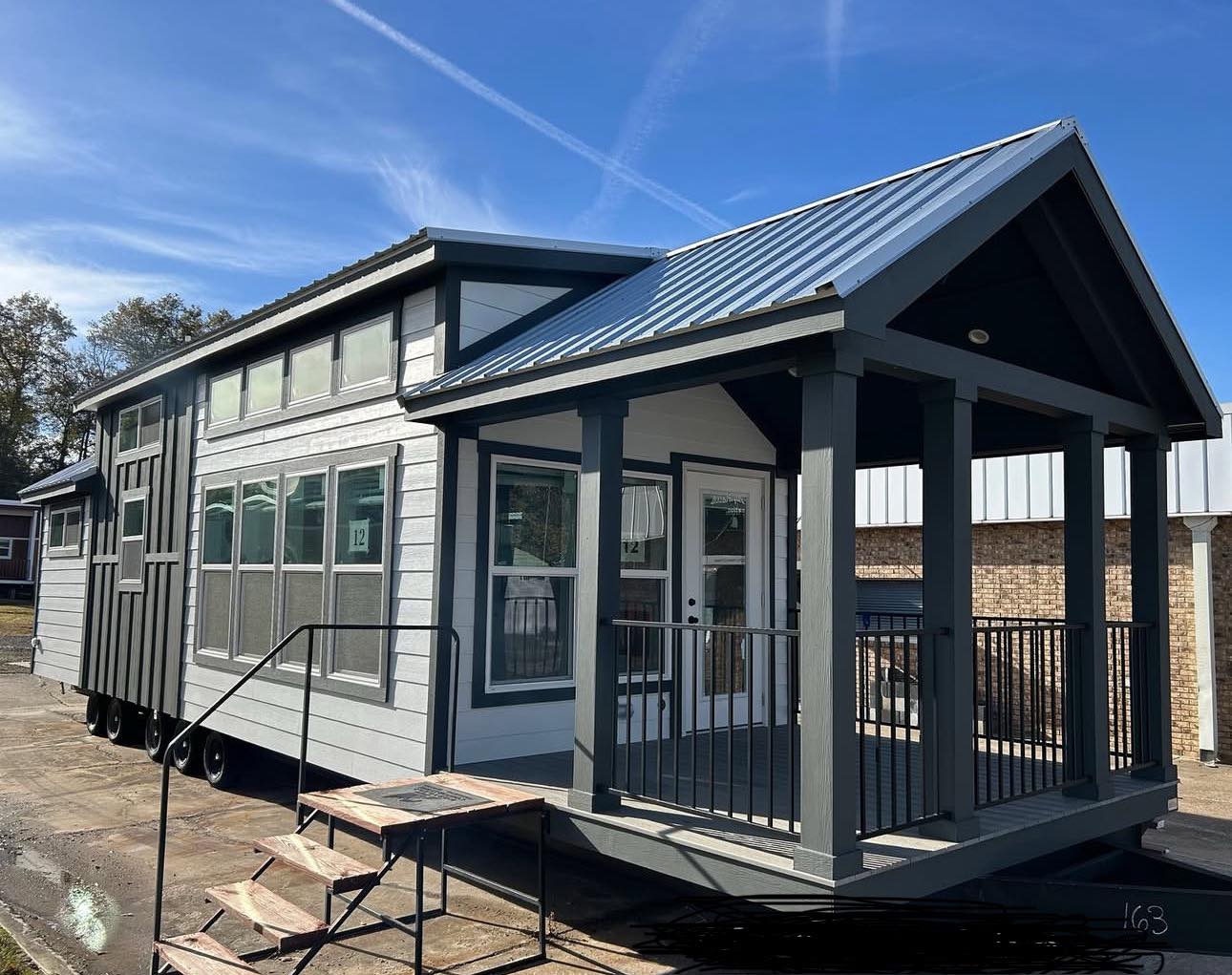A modern tiny house on wheels with gray siding and a metal roof. It features a covered porch with railings, large windows, and is parked on a sunny day.