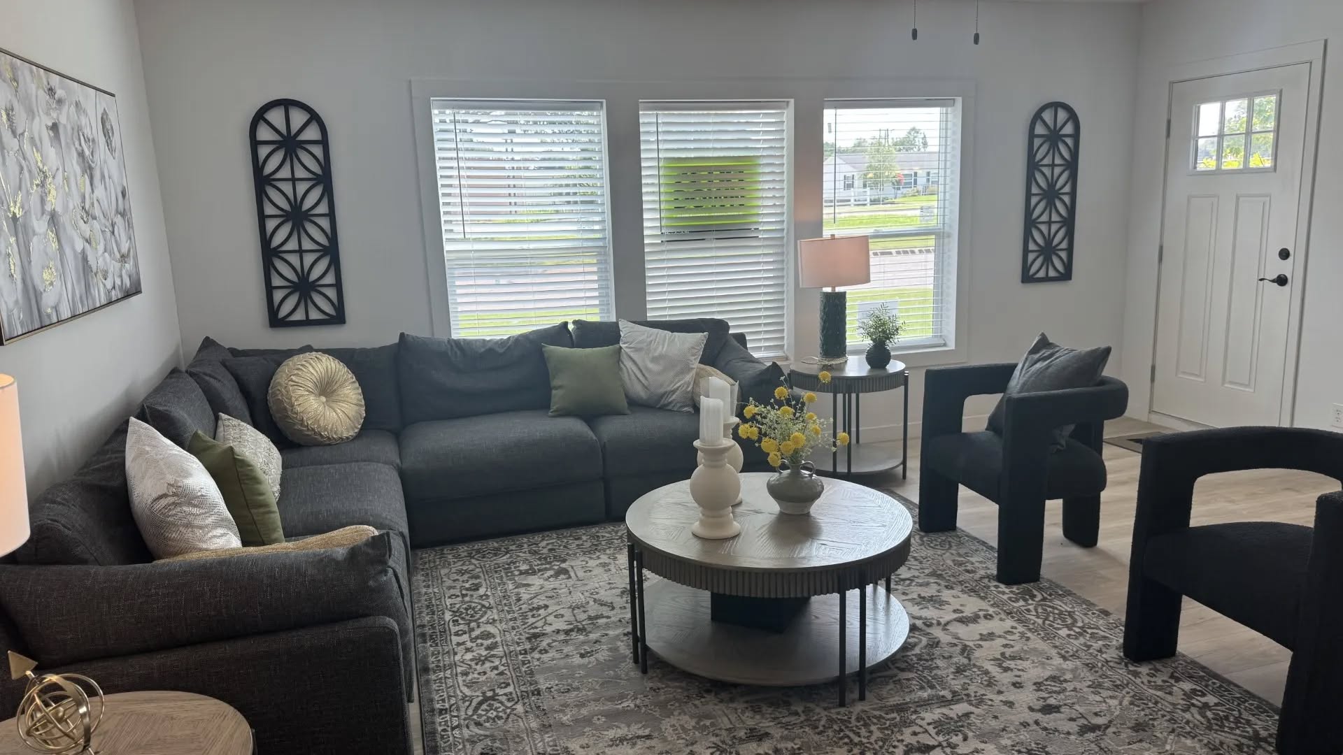 A cozy living room with a dark gray sectional sofa, decorative pillows, and two black chairs. A round coffee table holds vases and yellow flowers. Large, bright windows with blinds are in the background, flanked by geometric wall art. The ambiance is modern and inviting.
