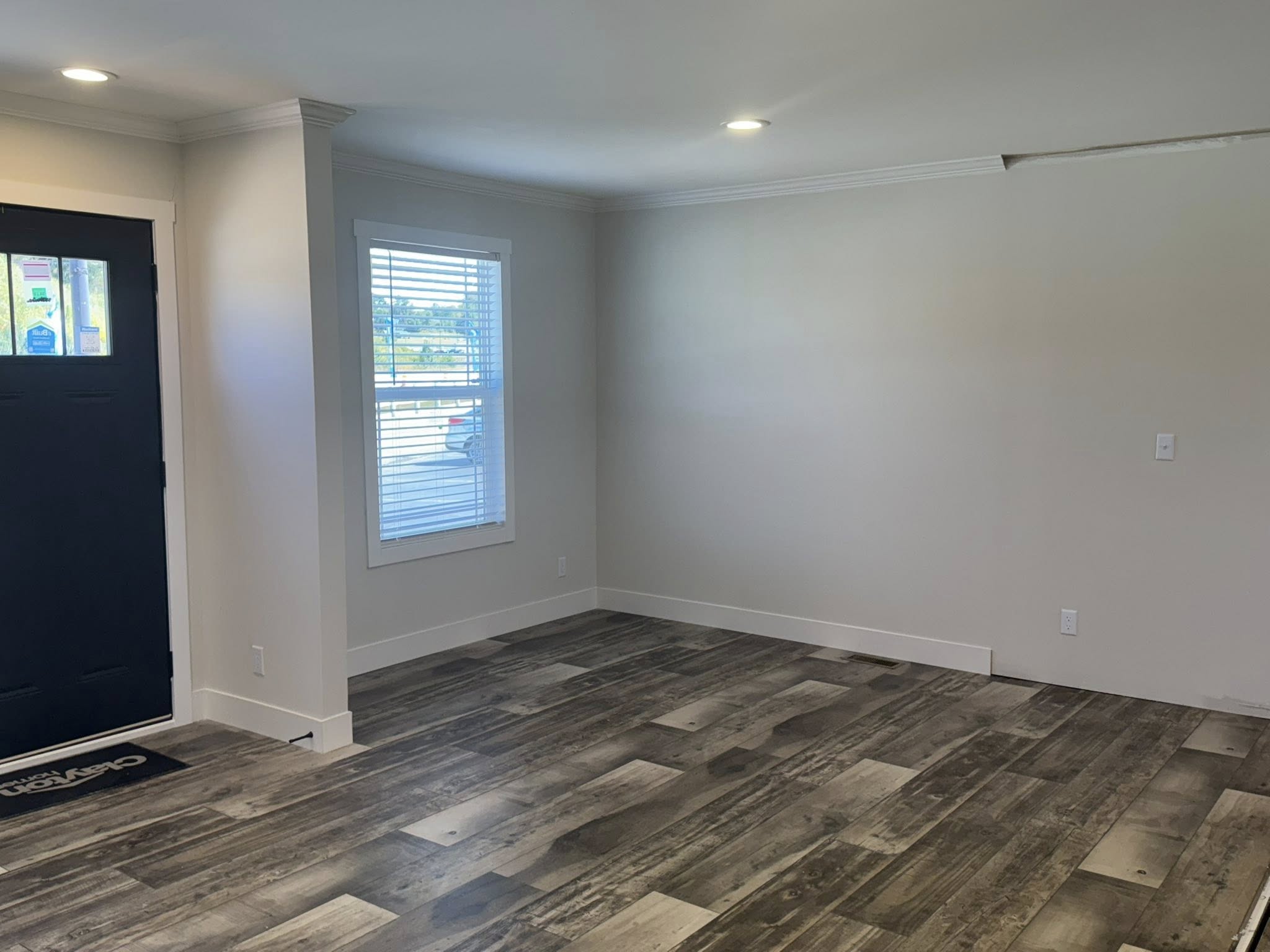 Spacious room with light gray walls, dark wooden floor, a closed black door, and a large window with blinds, creating a modern, airy atmosphere.
