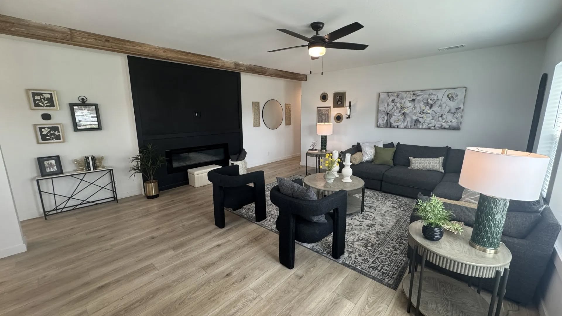 A modern living room with a gray sectional sofa, black chairs, and a patterned rug. There's a black accent wall, neutral decor, and warm lighting.