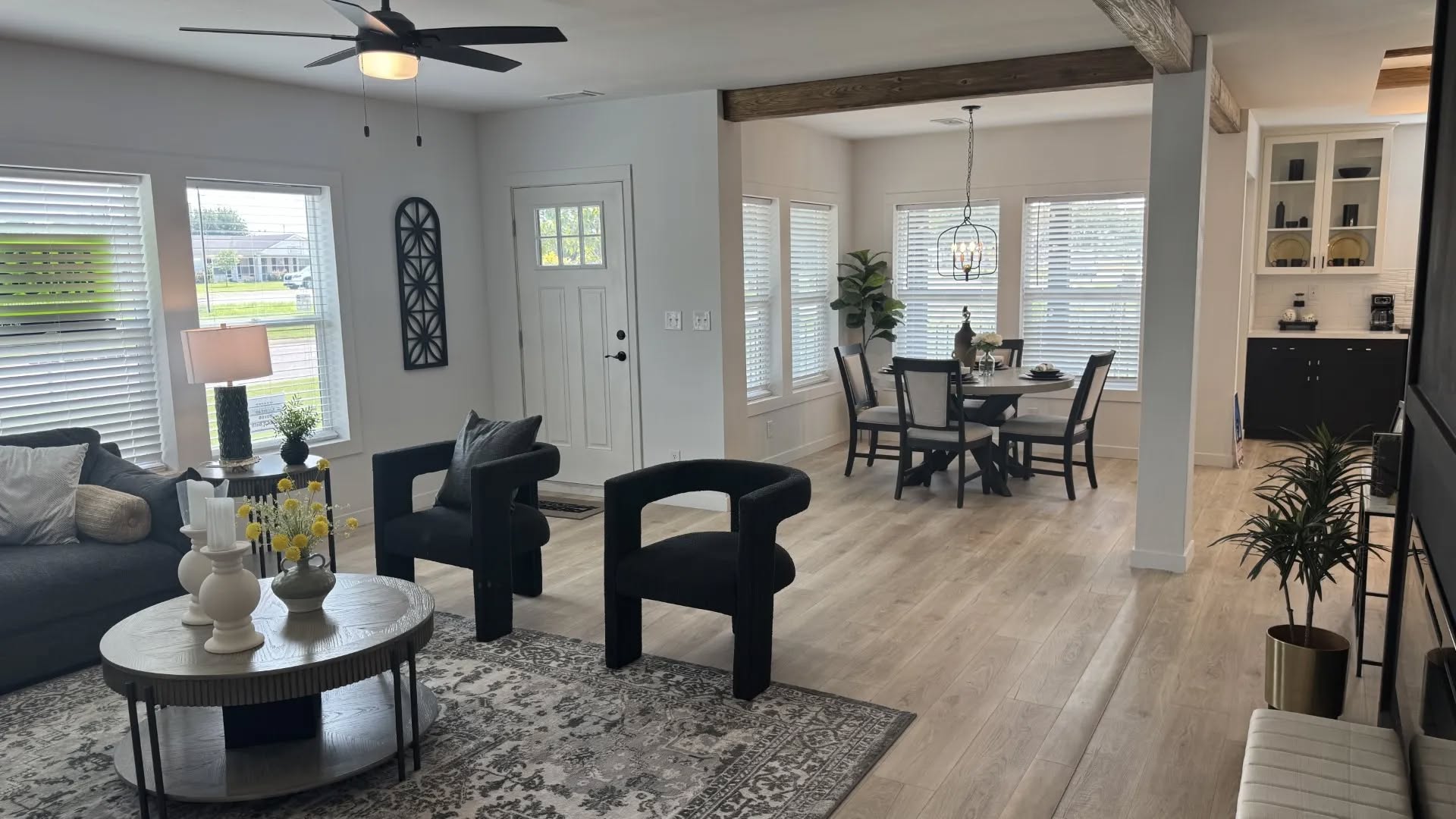 Modern living and dining area with light wood floors. The room has a gray sofa, round coffee table, black chairs, ceiling fan, and dining set. Large windows allow natural light, creating an airy feel.
