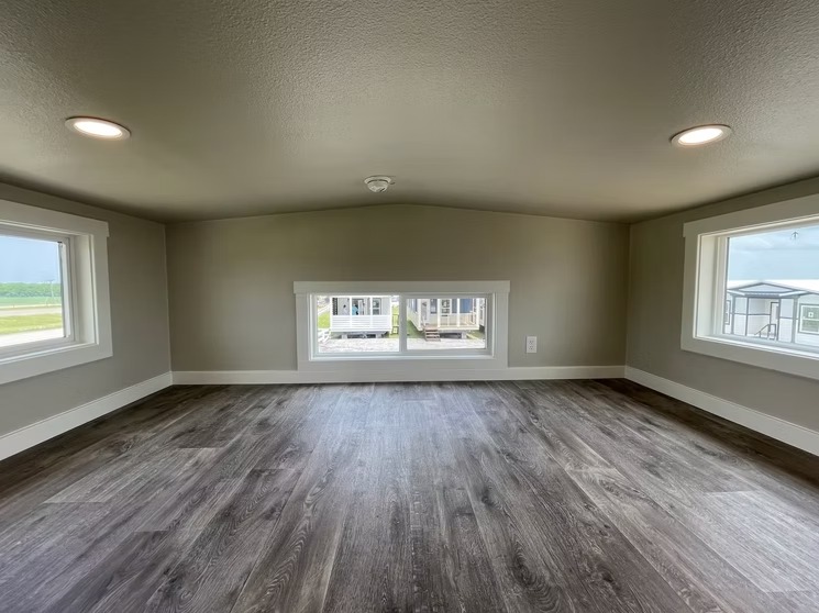 Small room with dark wooden flooring, gray walls, and three rectangular windows providing views of a grassy landscape and houses. Bright, cozy atmosphere.
