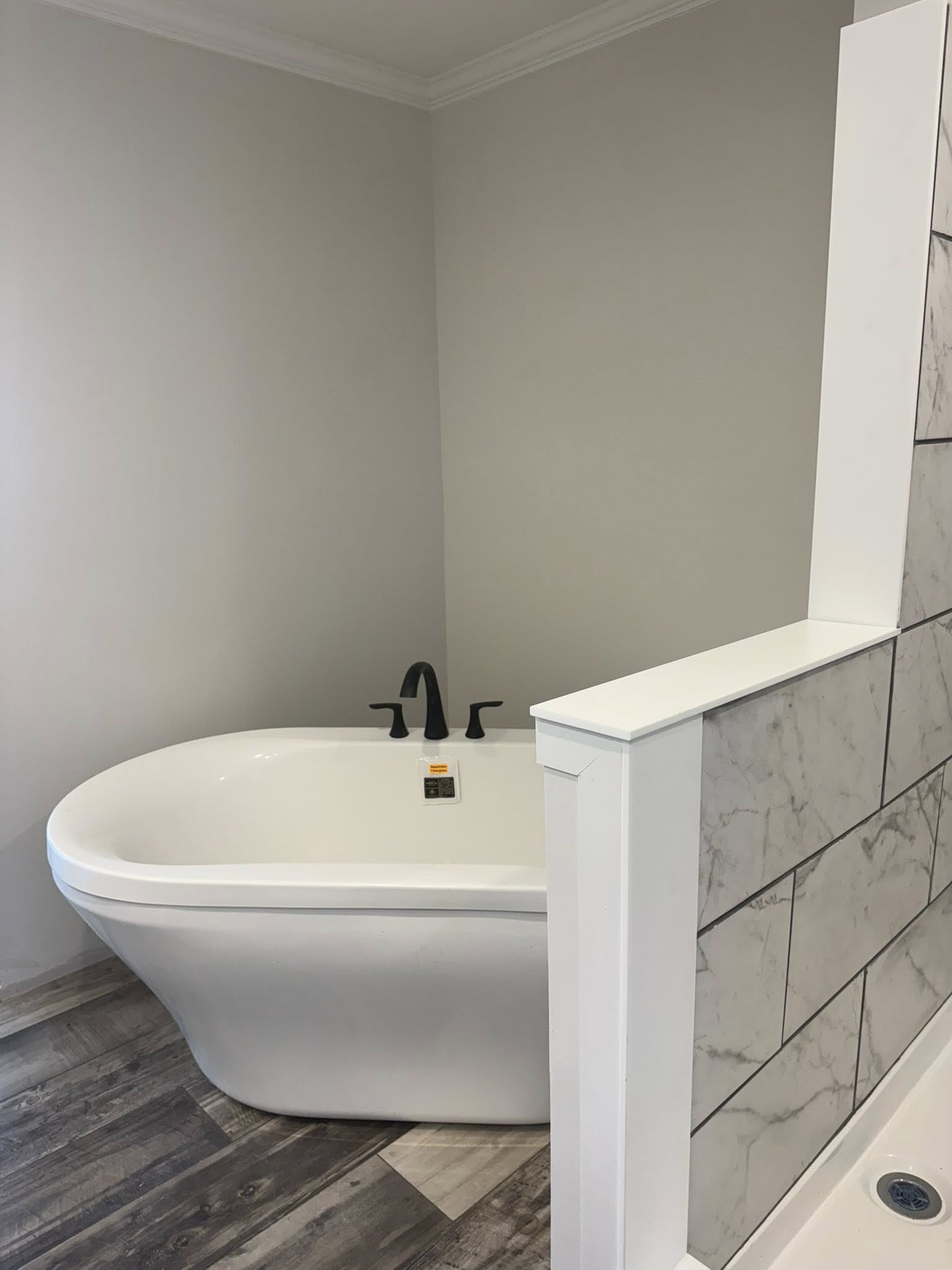 A modern bathroom with a white freestanding tub, black faucet, gray tiled half wall, and wood-look flooring. The atmosphere is clean and minimalist.