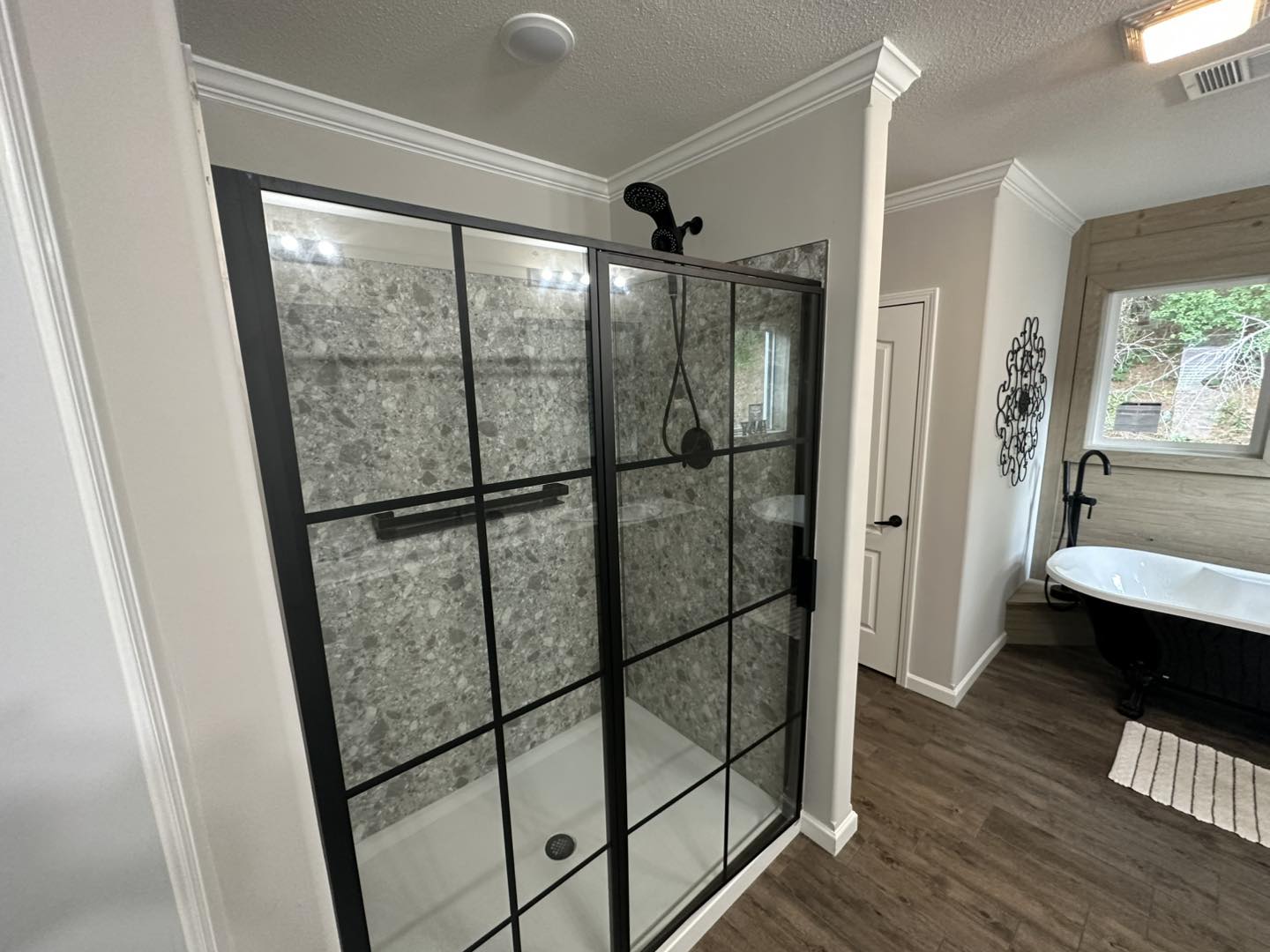 Modern bathroom with a black-framed glass shower, gray stone walls, and wood flooring. A white freestanding tub is by a window, creating a serene vibe.