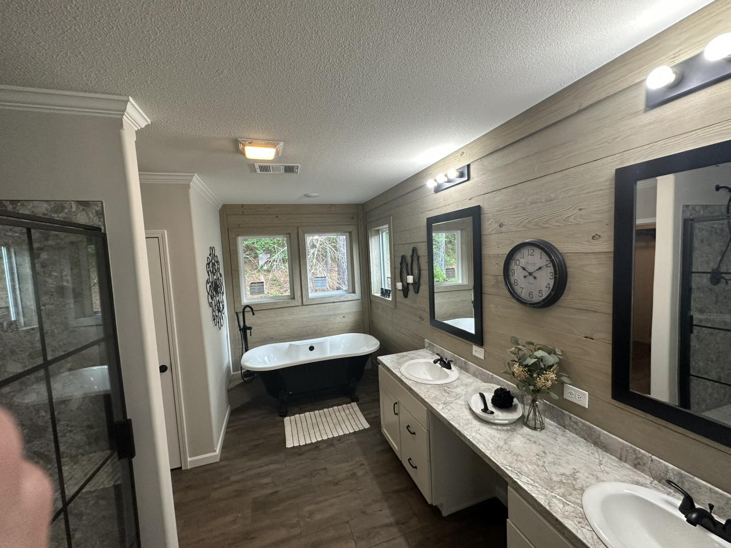 Bright bathroom with wood-paneled walls, featuring a black freestanding tub by large windows. Dual sinks on a marble counter, modern mirrors, and a wall clock.