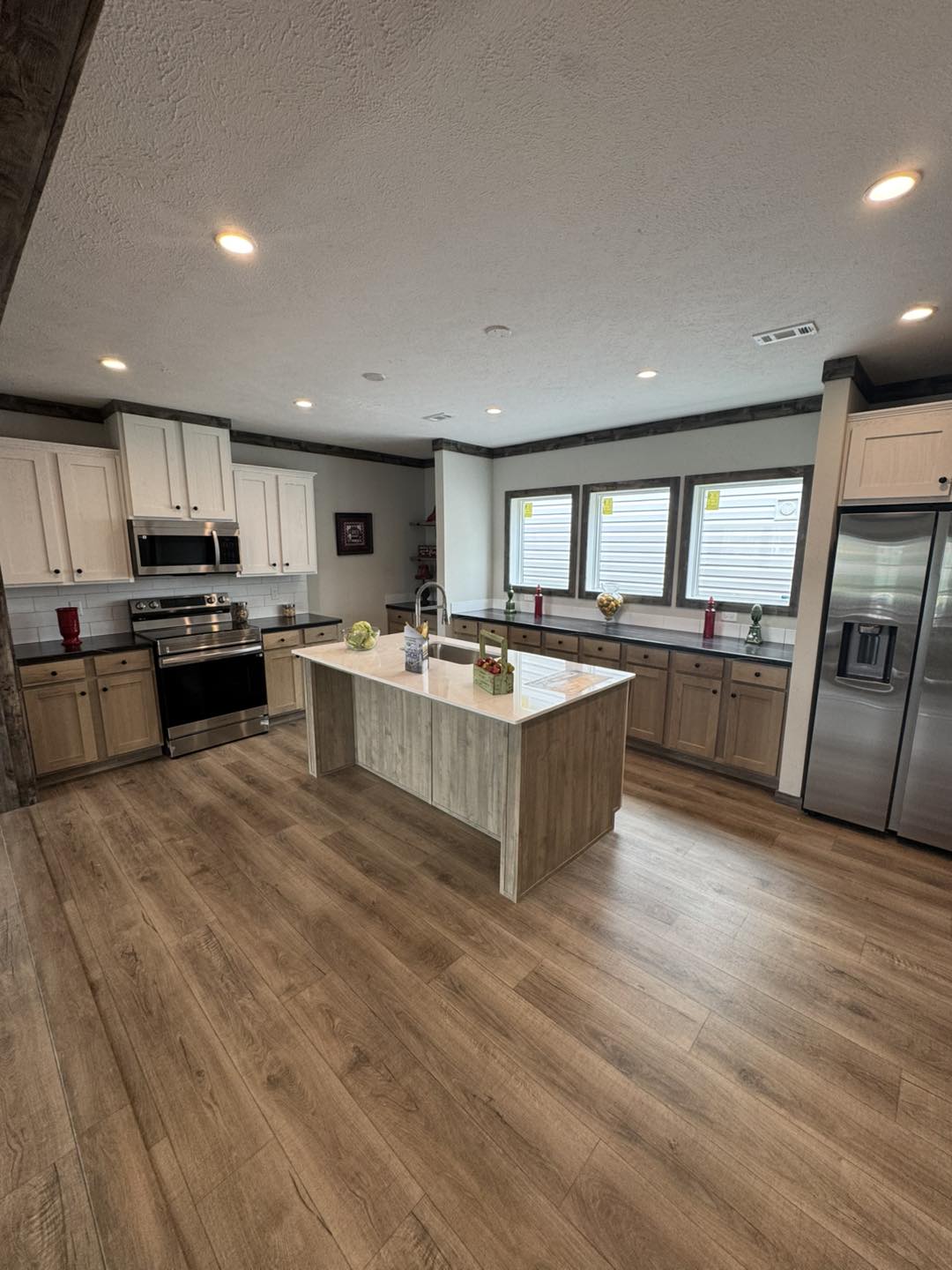 Spacious modern kitchen with wood flooring, centered island, stainless steel appliances, white and wood cabinets, and large windows with ample light.
