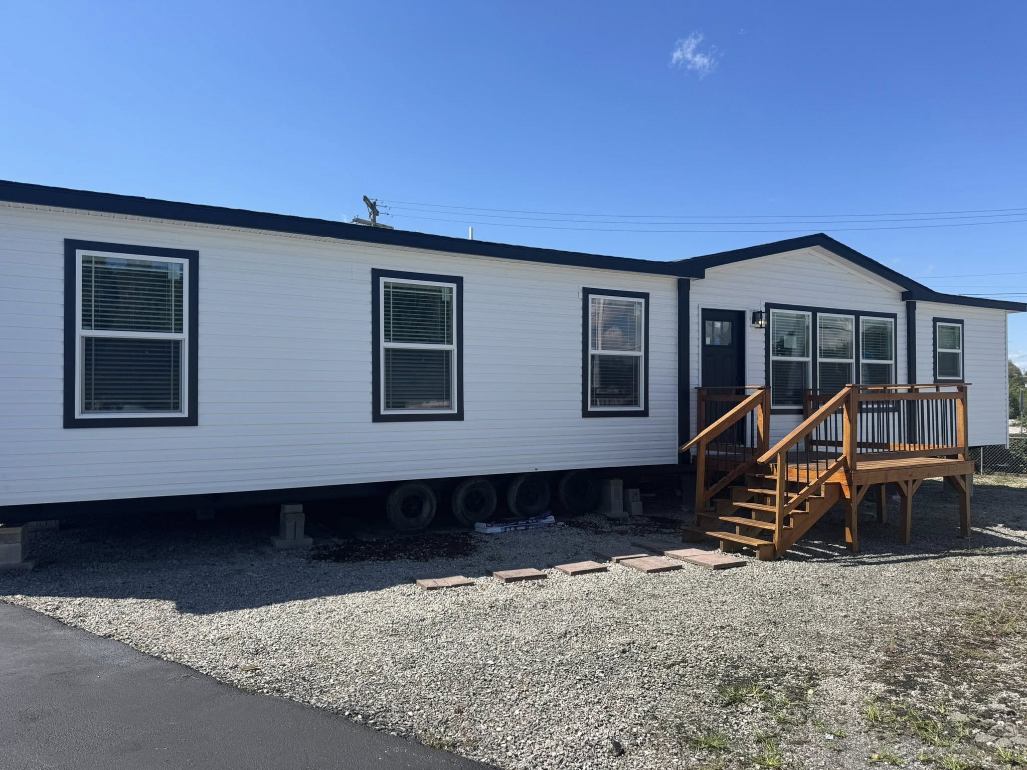 A white mobile home with black trim sits on gravel, featuring several windows and a wooden staircase leading to a small porch. The sky is clear and blue.