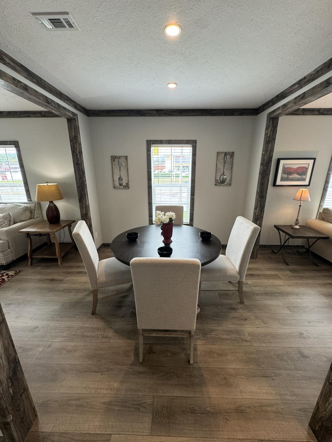 Cozy dining area with a round table and four beige chairs. Red vase with white flowers centers the table. Natural light from two side windows. Neutral decor.