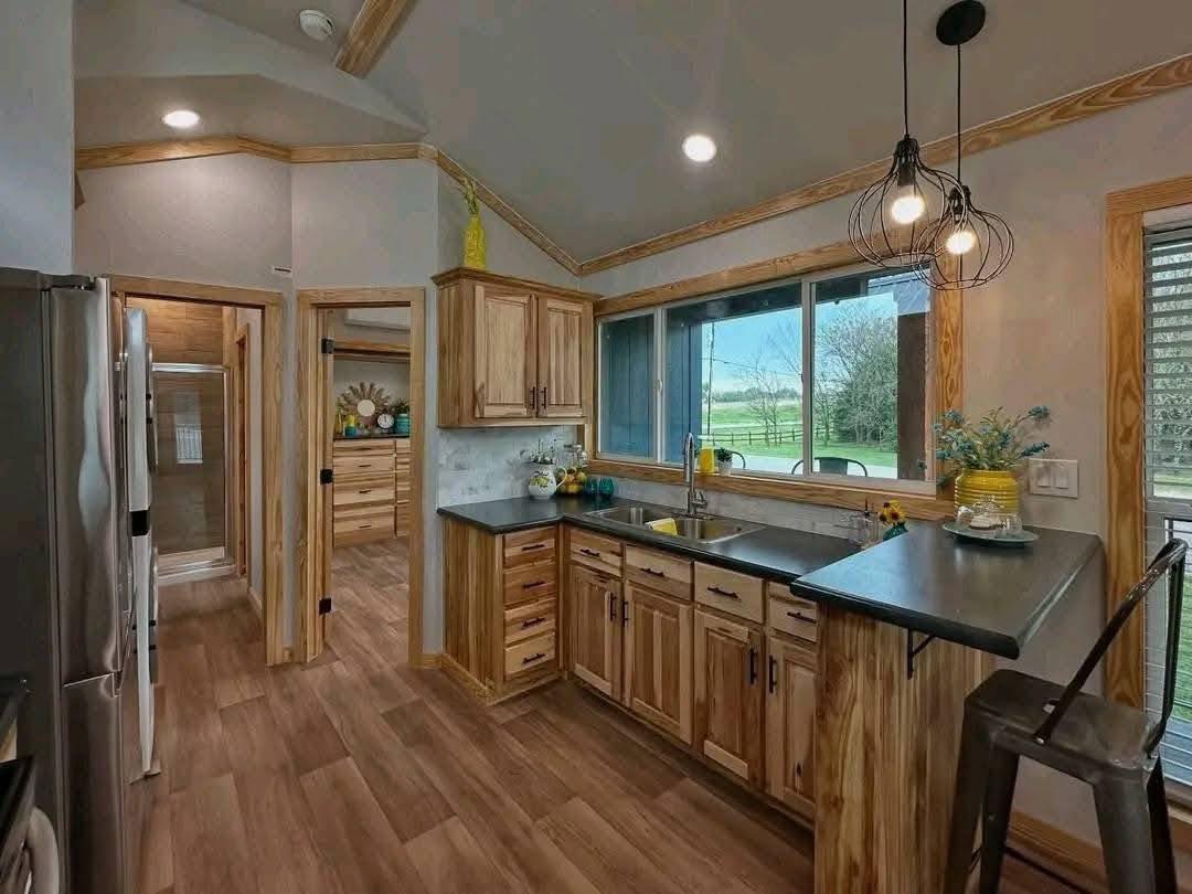 A cozy kitchen with wood cabinets and dark countertops. Large window offers a scenic view. Pendant lights hang above the counter, creating a welcoming atmosphere.