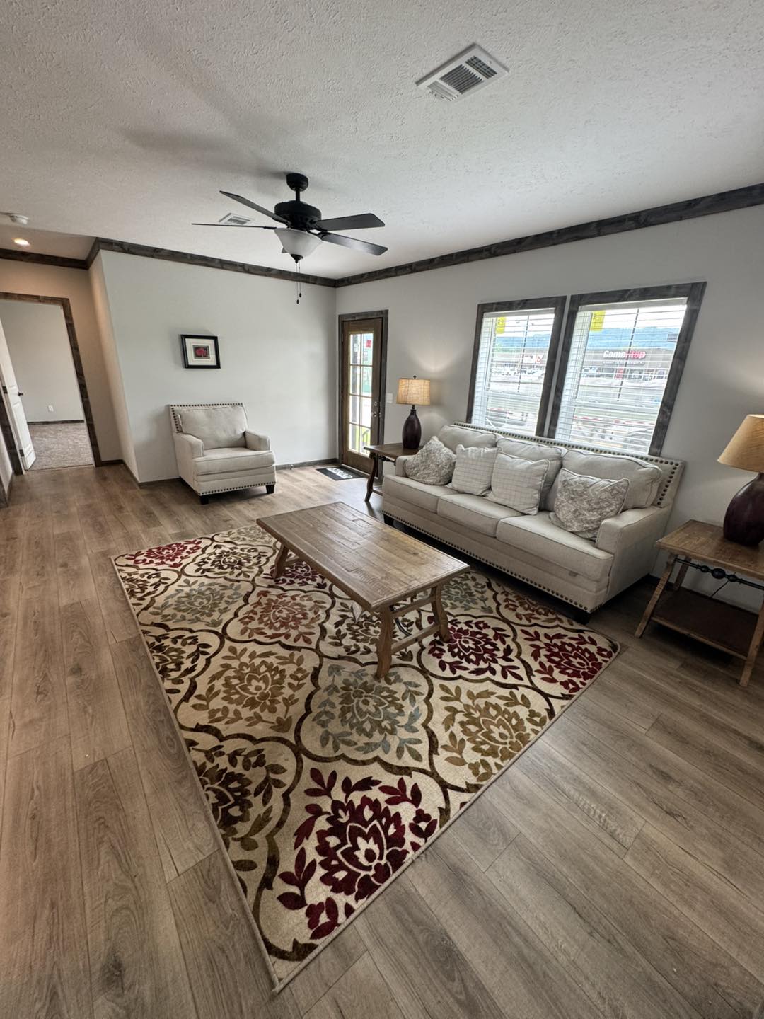 Cozy living room with beige sofa, armchair, and wooden coffee table on a floral-patterned rug. Large windows and neutral tones create a warm ambiance.