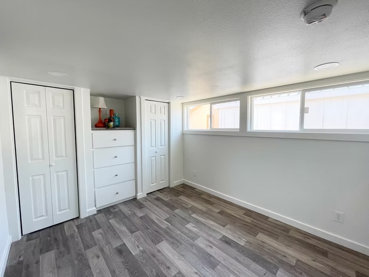 Small, bright basement room with large windows, white walls, and laminate flooring. Contains two closets and a chest of drawers with decor. Cozy ambiance.