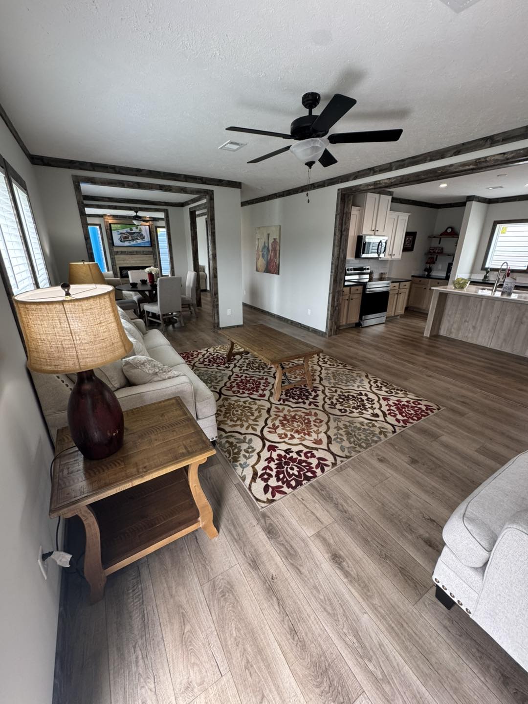 Spacious living area with wooden flooring, patterned rug, and neutral-toned furniture. Open layout includes a kitchen and dining space, creating an inviting atmosphere.
