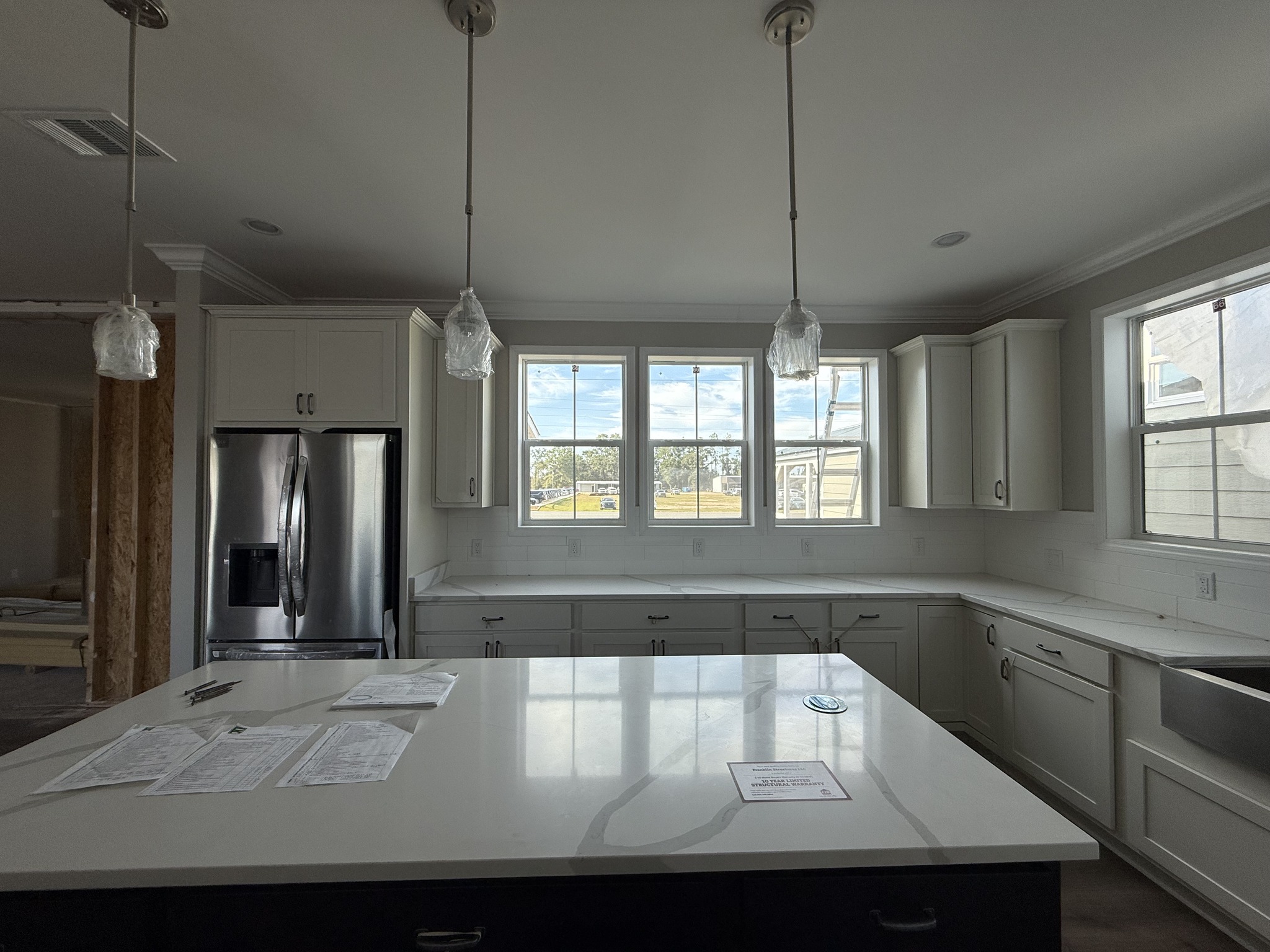 Modern kitchen with white cabinets, stainless steel fridge, and large island. Three pendant lights hang above. Papers lie on the island, and sunlight streams through multiple windows, creating a bright atmosphere.