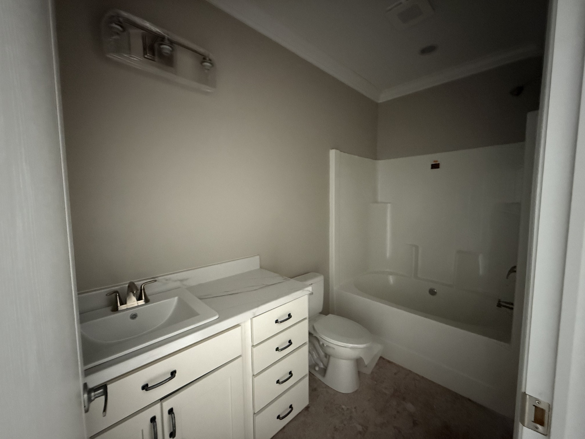 A small bathroom with beige walls features a white vanity with drawers, a sink, overhead light, and a bathtub. The atmosphere is simple and clean.