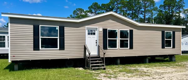 A beige manufactured home with a peaked roof sits on a grassy lot. It features white trim, black shutters, and small steps leading to a white front door.