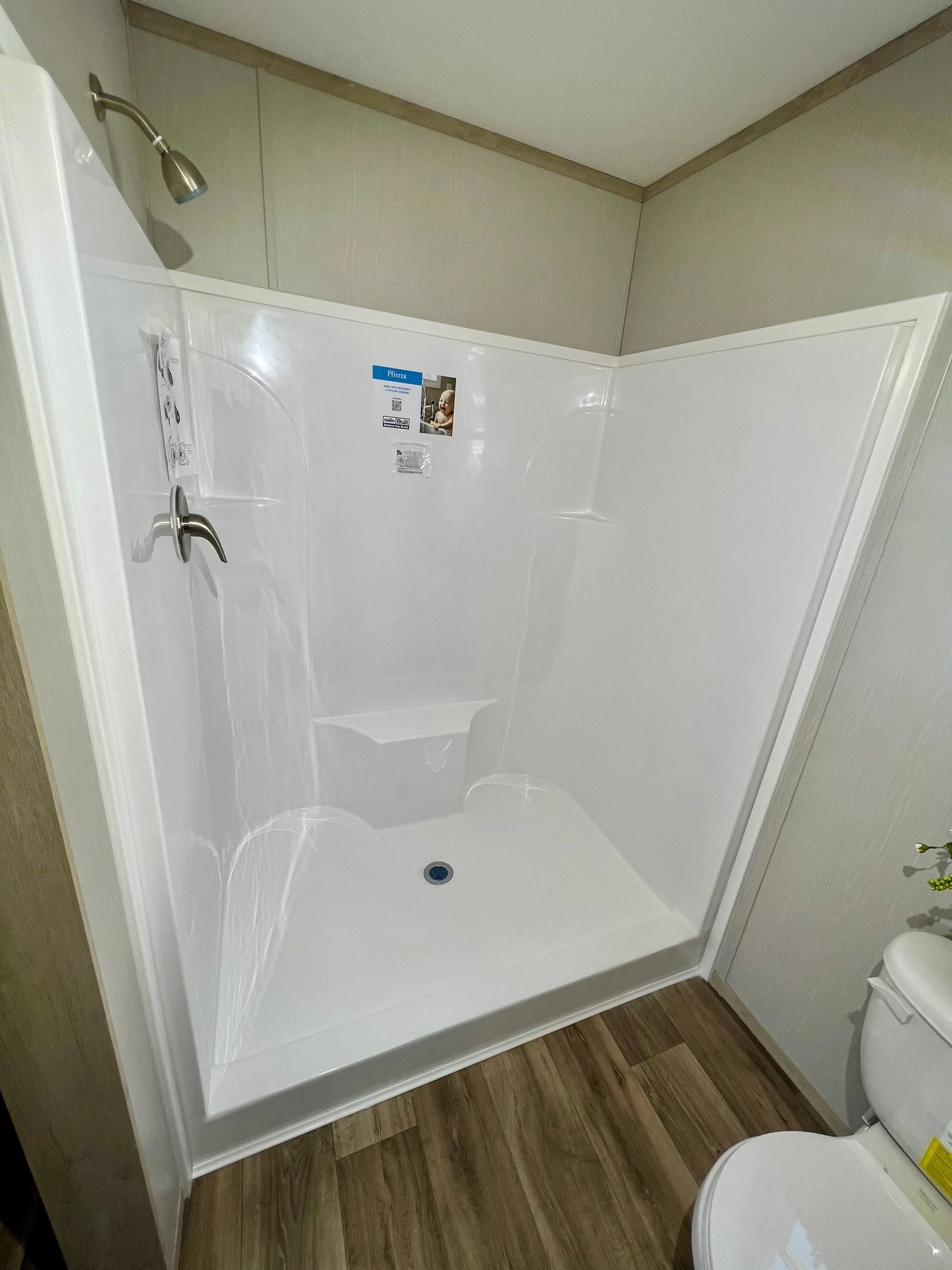 A clean, modern shower with white walls, silver fixtures, and a small built-in shelf inside. The floor is wood-patterned, and a white toilet is partially visible.