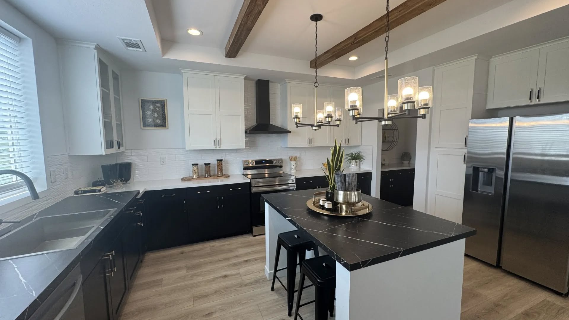 Modern kitchen with black and white cabinetry, stainless steel appliances, and a black island. Wood beams and a chandelier add warmth and elegance.