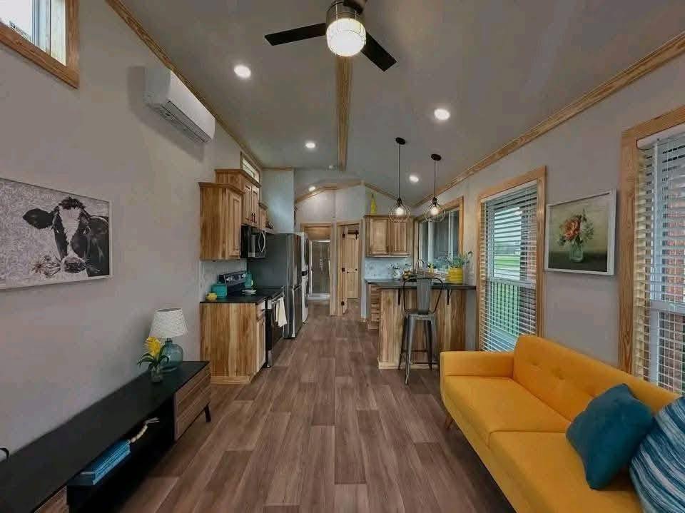 Bright, modern living space with wood flooring, a yellow sofa, and blue cushions. A cow painting and sunflowers add charm. Stylish kitchen with bar stools. Cozy and inviting.