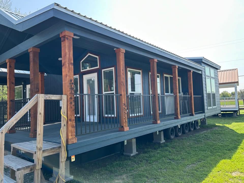 A modern tiny house on wheels with dark exterior, wooden pillars, and a small staircase. Sunlit windows and green grass convey a warm, inviting feel.